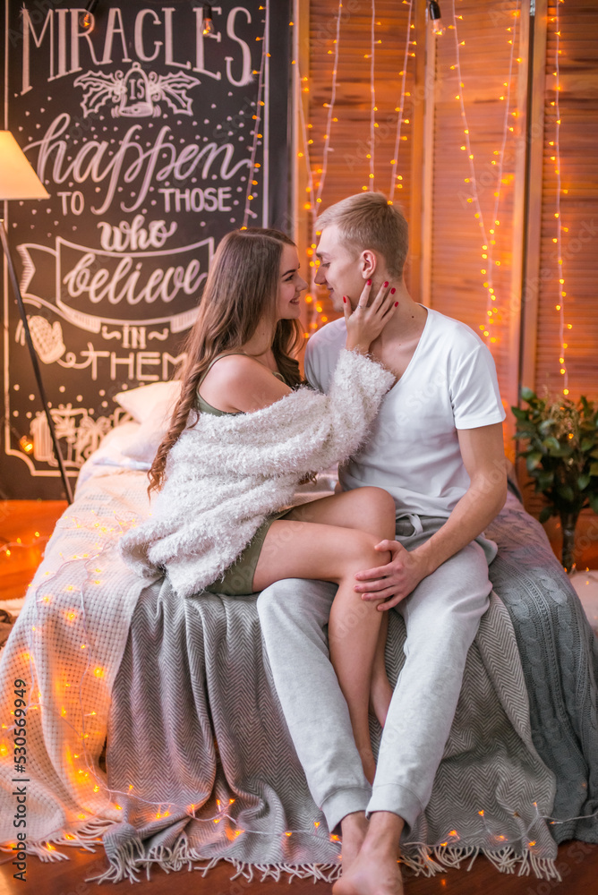 Obraz premium A young and attractive couple, a guy and a girl are sitting and hugging on a bed in a room decorated for Christmas with garlands. Christmas mood