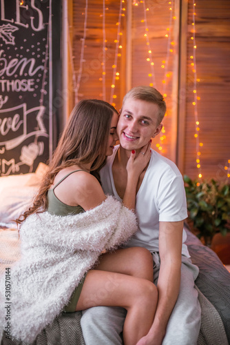 A young and attractive couple, a guy and a girl are sitting and hugging on a bed in a room decorated for Christmas with garlands. Christmas mood © capable97