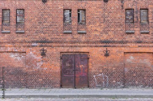 Wall of an old red brick factory building with narrow windows and metal gates