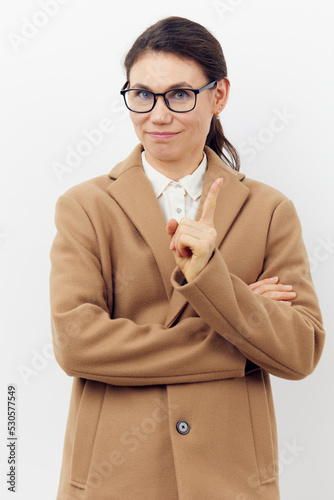 a sweet, pleasant woman with glasses is standing in a stylish beige coat on a light background and pointing her finger up