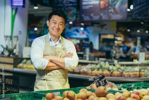 portrait of a happy handsome Asian salesman. Vegetables and fruits at the market or grocery store. Seller. a man in an apron looks at the camera and smiles farmer, small business owner greengrocer photo