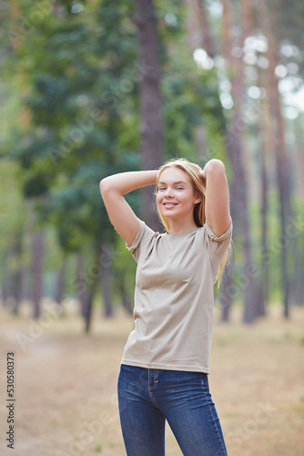 Blonde lady with natural makeup posing in a pine forest. Gorgeous blue eyed caucasian woman in beige t-shirt , enjoying summer vacation.