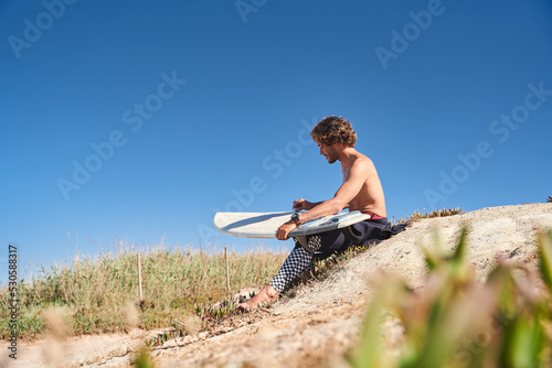 Shirtless surfer waxing white surfboard with pattern while sitting at the beach