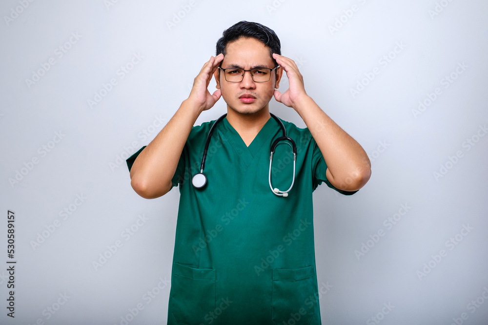 Stressed Asian young doctor or male nurse in green uniform with stethoscope having headache Stressed Asian young doctor or male nurse in green uniform with stethoscope having headache