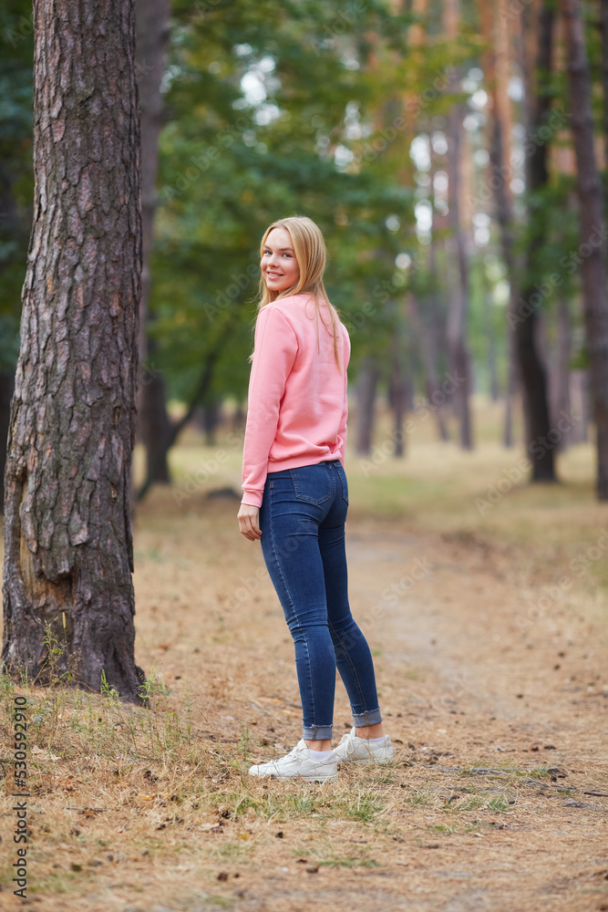 Blue-eyed blonde in a pink hoodie walks in a pine forest. Portrait of a joyful young woman enjoying in autumn park.