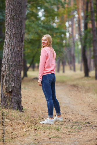 Blue-eyed blonde in a pink hoodie walks in a pine forest. Portrait of a joyful young woman enjoying in autumn park.