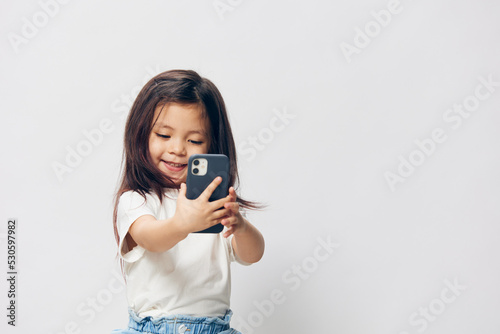 a cute, little girl of preschool age in a white T-shirt stands on a light background and holds a fashionable smartphone in her hands looking enthusiastically into it