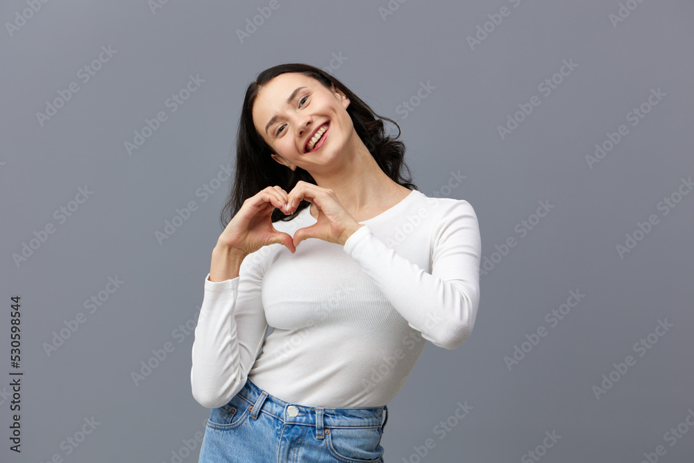 an attractive woman stands on a gray background in a white T-shirt, happily made a heart out of her hands an attractive woman stands on a gray background in a white T-shirt, happily made a heart out of her hands