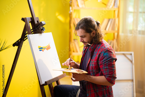 A beautiful man with a beard and a manbun is concentrating on the art he is painting with lots of colours on his canvas photo