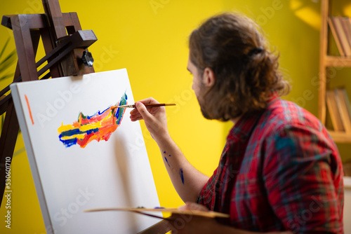 A painter is looking directly into the camera and smiling as he is holding up his art palette and sitting in a chair in his art atelier