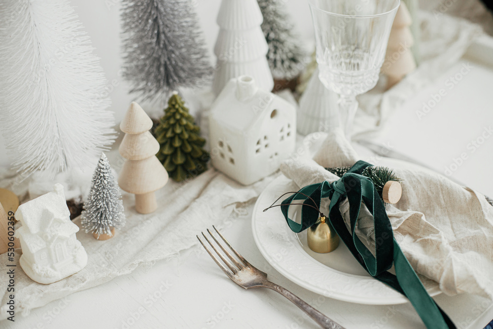 Christmas table setting. Stylish napkin with ribbon and bell on plate, vintage cutlery, modern festive christmas trees and houses on white rustic table. Holiday arrangement of table Christmas table setting. Stylish napkin with ribbon and bell on plate, vintage cutlery, modern festive christmas trees and houses on white rustic table. Holiday arrangement of table