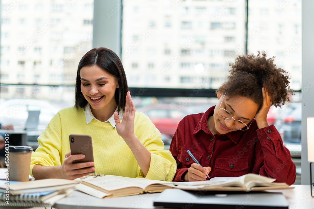 Waist up portrait view of the caucasian overjoyed woman gesturing while using her smartphone. Her multiracial classmate studying nearby. Library concept Waist up portrait view of the caucasian overjoyed woman gesturing while using her smartphone. Her multiracial classmate studying nearby. Library concept