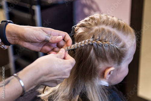 Hair salon, Hairdresser makes hairdo for baby in barber shop close up. Barber woman make fashionable braids hairstyle for cute little blond girl child in modern barbershop. Concept hairstyle, beauty