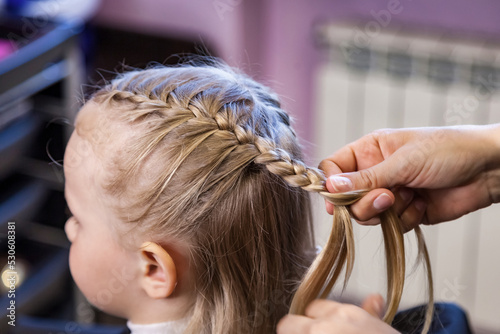 Hair salon, Barber woman make fashionable braids hairstyle for cute little blond girl child in modern barbershop. Hairdresser makes hairdo for baby in barber shop close up. Concept hairstyle, beauty