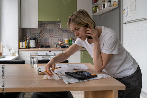 Mature woman talks on smartphone calling different hotlines. Middle aged female with upset expression tries to solve problems with unpaid bills checking papers on table. Utility debt and rates concept photo
