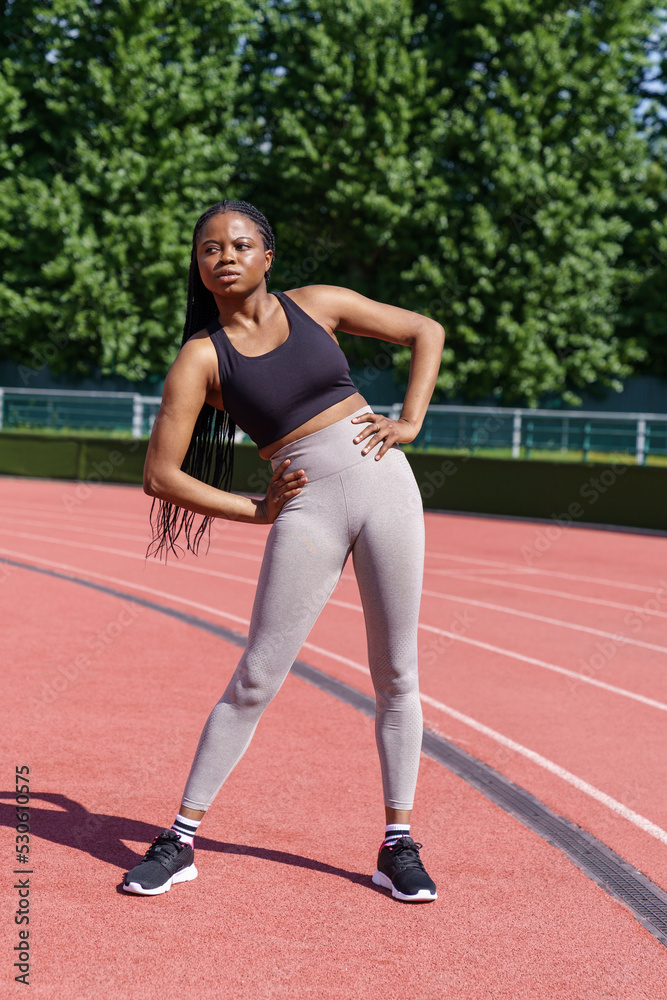 Black young sportswoman with long braids enjoys warming up before intensive workout on big city stadium on sunny day. Athletic African American female does pelvic tilts to sides standing on red track