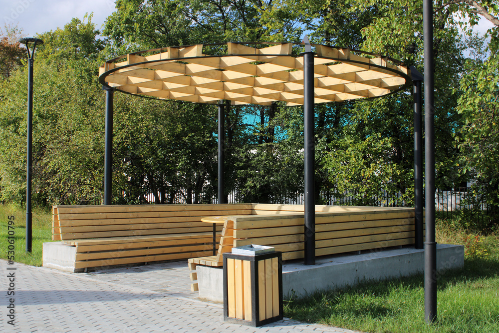 A light wooden gazebo with a bench, a table and an unusual hutch in a city park. Urban architecture
