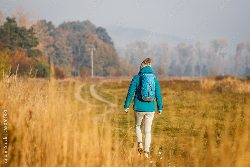 Woman with backpack hiking on footpath at cold morning. Fall exploration outdoors in rural scenery