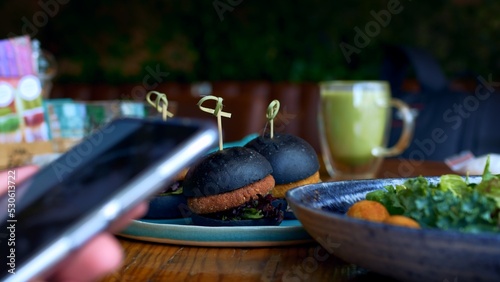 At a restaurant table  a man sits on his phone and looks at the screen by swiping his fingers over it. On the table are salad dishes and three hamburgers with various fillings and black buns.