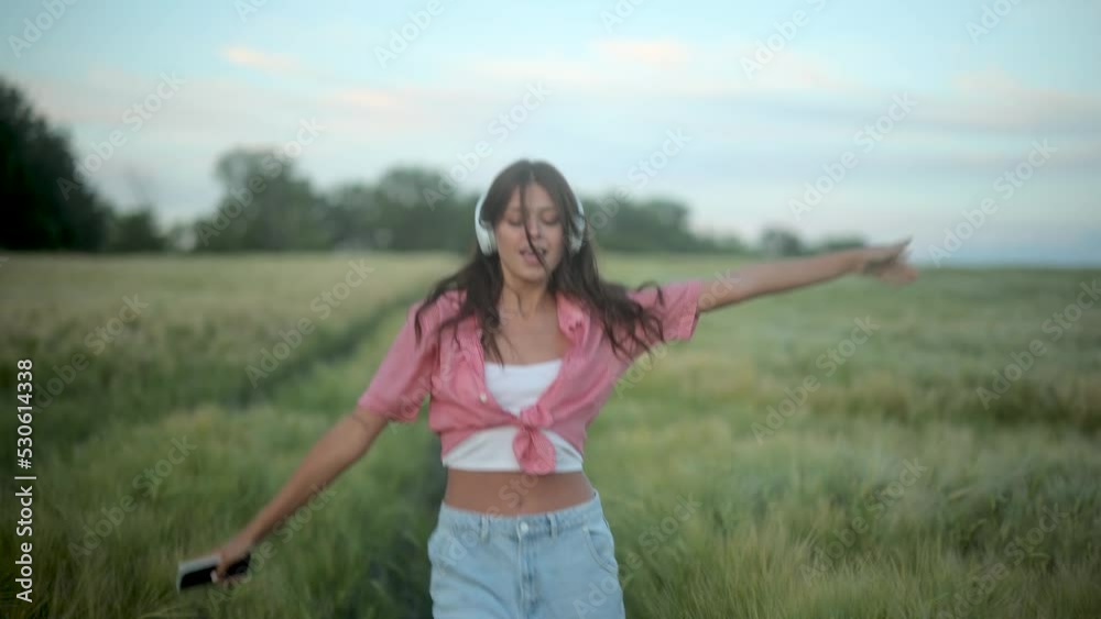 Young beautiful woman in jeans and checkered shirt listens to music and dances while walking in the field during sunset Young beautiful woman in jeans and checkered shirt listens to music and dances while walking in the field during sunset