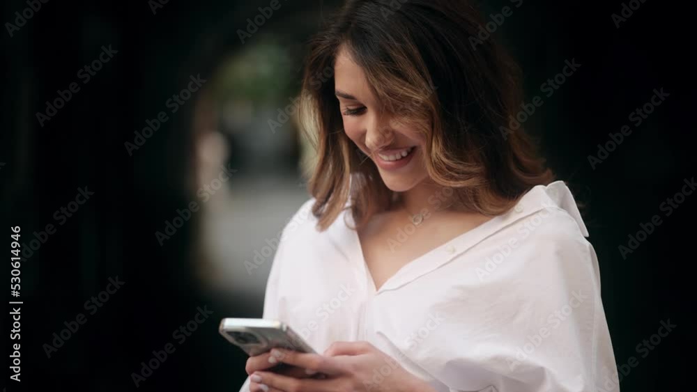 Happy woman messages via mobile phone application uses free internet connection wears white shirt smiles gladfully stands on city street Happy woman messages via mobile phone application uses free internet connection wears white shirt smiles gladfully stands on city street