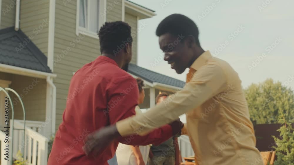 Medium shot of two happy grown up Black men and their families gathering together for dinner in yard outside their house on summer day, greeting each other with joy Medium shot of two happy grown up Black men and their families gathering together for dinner in yard outside their house on summer day, greeting each other with joy