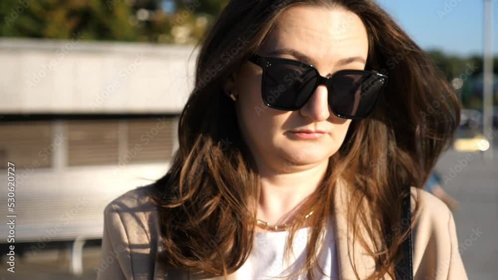 Portrait of woman in sunglasses with displeased facial expression. Serious brunette girl looks into camera at city street. Unhappy irritated businesswoman. Concept of sorrowful or annoyed emotion Portrait of woman in sunglasses with displeased facial expression. Serious brunette girl looks into camera at city street. Unhappy irritated businesswoman. Concept of sorrowful or annoyed emotion
