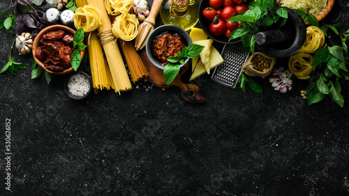 Dry spaghetti and ingredients for their preparation: basil, parmesan, pine nuts, tomatoes, spices. Italian traditional cuisine. On a black stone background background.