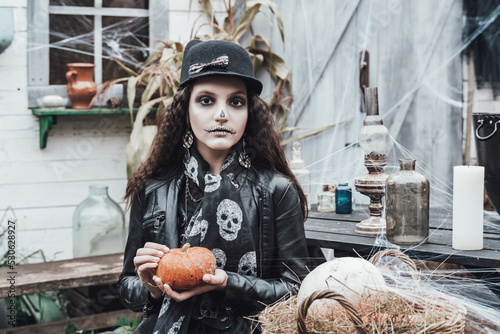 Beautiful scary little girl celebrating halloween. Terrifying black, white half-face makeup,witch costume, stylish image. Horror, fun at children's party in barn on street. Hat, jacket, pumpkin