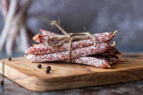 Italian sausage snack. Delicious dry sausage with a bow on a wooden board, black concrete background and a pile of dry sausage behind it.