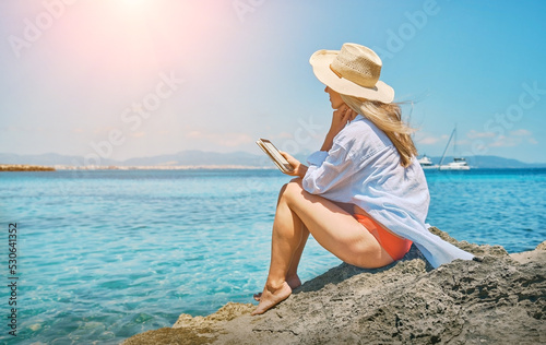 Fashion pretty woman outdoors lifestyle watching, reading on tablet ebook on the beach in summer day. Wearing wide brimmed hat, Sunbating with uv protection. Concept of beach vacation.