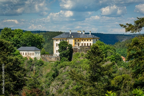 Obraz na płótnie Rabstejn nad Strelou, Czech Republic - June 12 2022: View of the white and yellow building, a castle, standing on a rock. Green trees in the foreground. Blue sky with clouds. Sunny summer day. w salonie