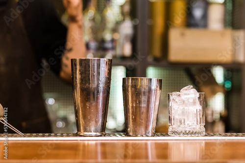 There is a shaker and a glass of ice on the bar counter. The glass is cooled to serve a cocktail