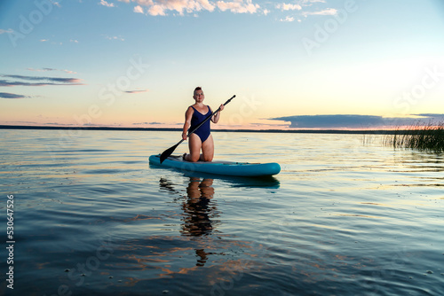 a woman in a closed swimsuit on her knees on a SUP board with an oar floats on the water against the background of the sunset sky.