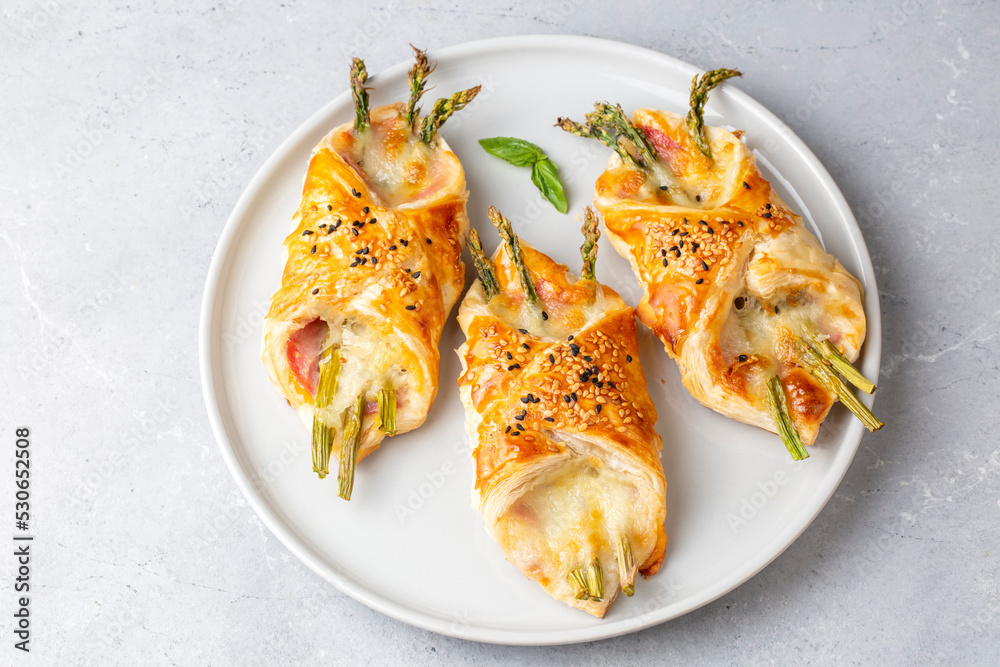 Baked green asparagus with ham and cheese in puff pastry sprinkled with sesame seeds and green basil leaves.