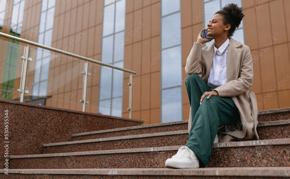 african american young woman talking on the phone while sitting on the steps against the backdrop of a business center