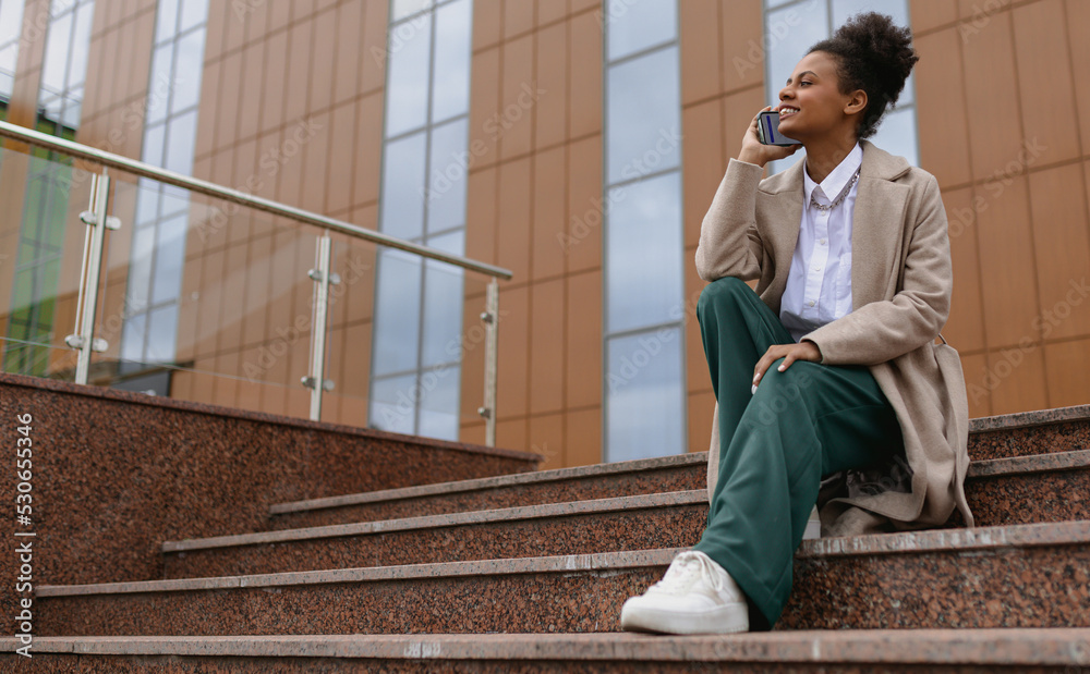 Fototapeta premium african american young woman talking on the phone while sitting on the steps against the backdrop of a business center