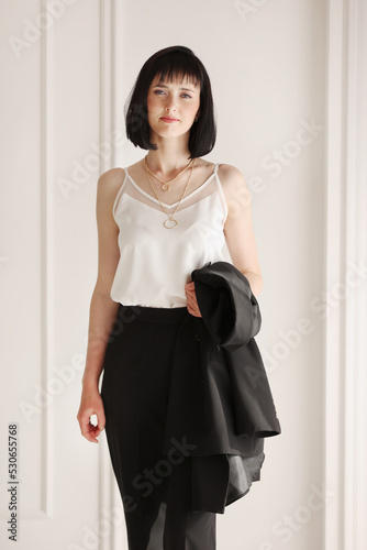 Confident young brunette business woman looking at camera on white wall background. Studio portrait of successful friendly female with short hair in black suit and white blouse with jacket on shoulder