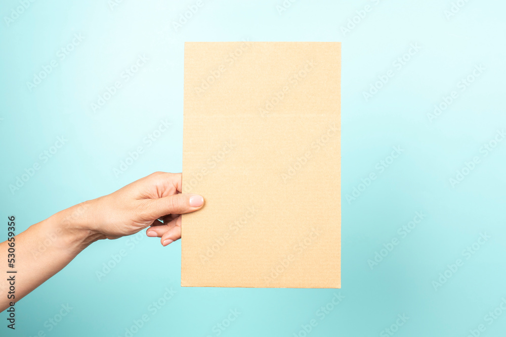 Woman hand holding blank rectangular sheet of brown cardboard on light blue background Woman hand holding blank rectangular sheet of brown cardboard on light blue background