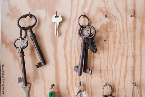 Various vintage and modern door keys hang on hooks on a wooden board. 