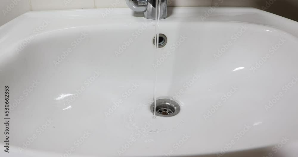 Stream of water falling from the metal faucet and running down the drain into a white ceramic sink, clean drinking water