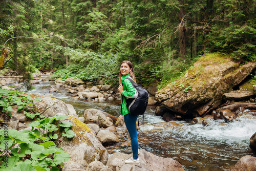 Traveler hiker with backpack enjoys landscape of mountain river in Carpathian forest. Young woman walks on rocks