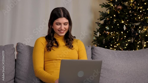 Smiling young woman uses laptop online chatting with best friends on background Christmas tree. Positive beautiful female distance works for computer typing text on keyboard sitting on couch at home