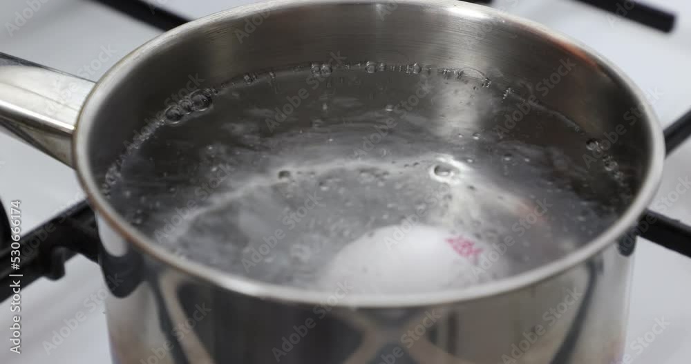 Close-up of the introduction of four white eggs on a spoon into the clean and transparent boiling water, black grill, white gas stove, metal pan. Home food concept Close-up of the introduction of four white eggs on a spoon into the clean and transparent boiling water, black grill, white gas stove, metal pan. Home food concept