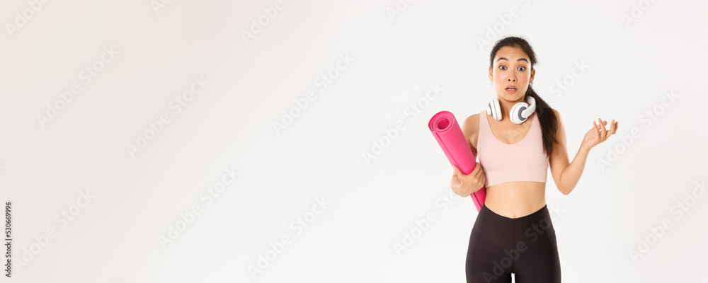 Sport, wellbeing and active lifestyle concept. Confused and shocked asian fitness girl, cant understand why gym closed, holding rubber mat and looking surprised at camera, white background - obrazy, fototapety, plakaty 