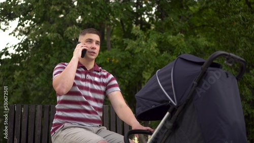 A young father is talking on the phone and rocking the stroller. A man with a stroller on a bench in the park.