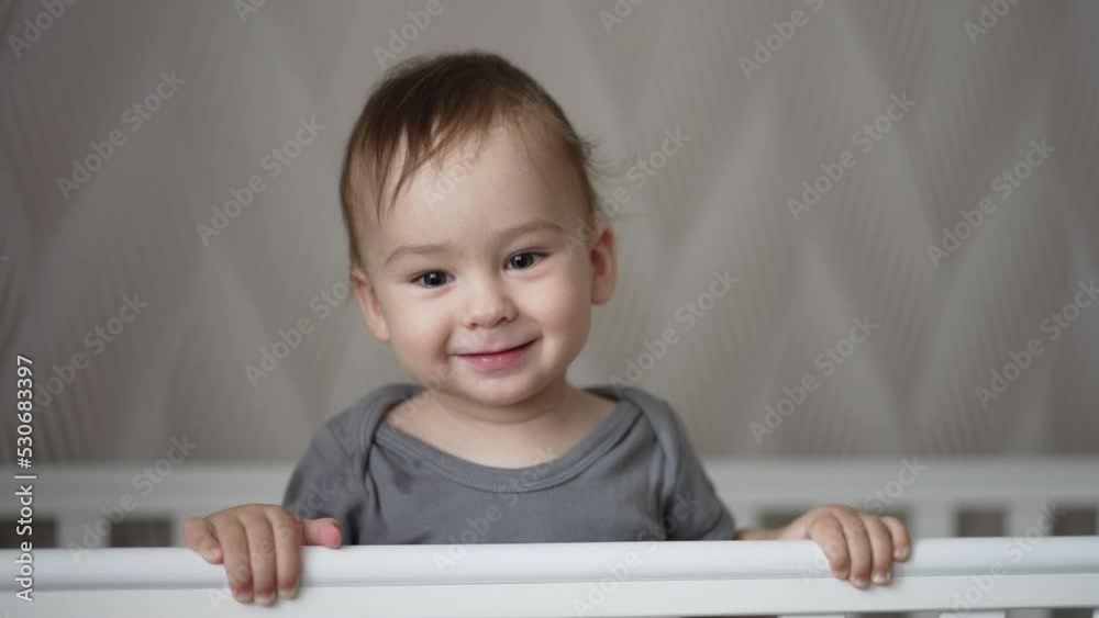 Adorably smiling baby boy and showing his two lower teeth. Sweet little toddler indoors in white baby cot. Close up. Blurred backdrop. Adorably smiling baby boy and showing his two lower teeth. Sweet little toddler indoors in white baby cot. Close up. Blurred backdrop.