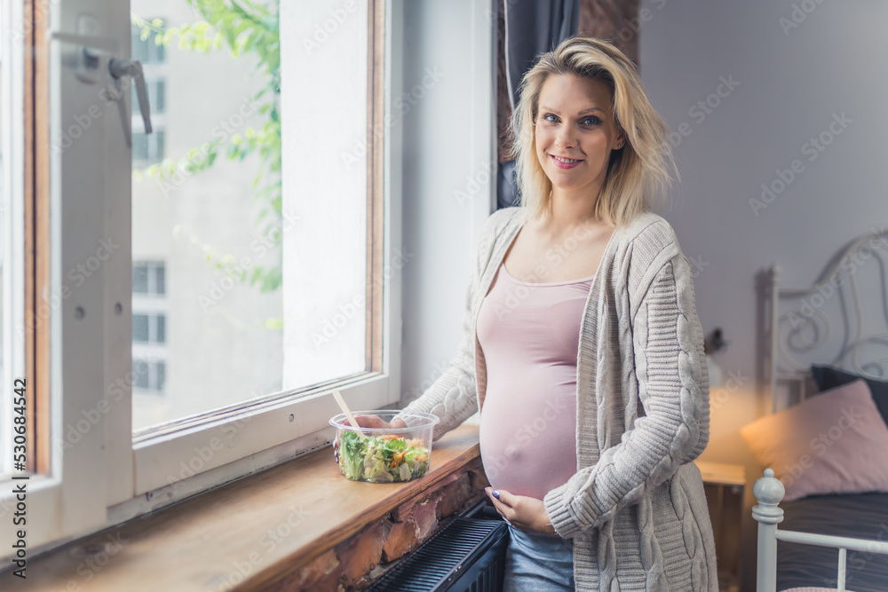 Healthy eating during pregnancy. Young caucasian pregnant woman standing near the window with a nutritious, rich in vitamins salad meal for growing a healthy baby. High quality photo