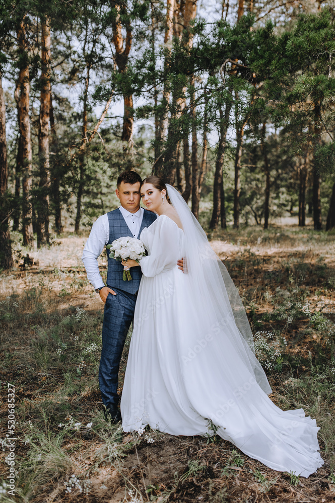 Beautiful newlyweds are hugging in the forest. Wedding portrait, photo of a stylish groom and a cute brunette bride in a white long dress with a bouquet of roses. Beautiful newlyweds are hugging in the forest. Wedding portrait, photo of a stylish groom and a cute brunette bride in a white long dress with a bouquet of roses.