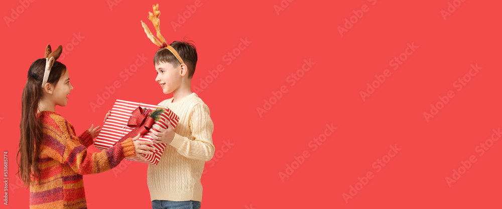 Fototapeta premium Funny little children with reindeer horns and Christmas gift on red background with space for text